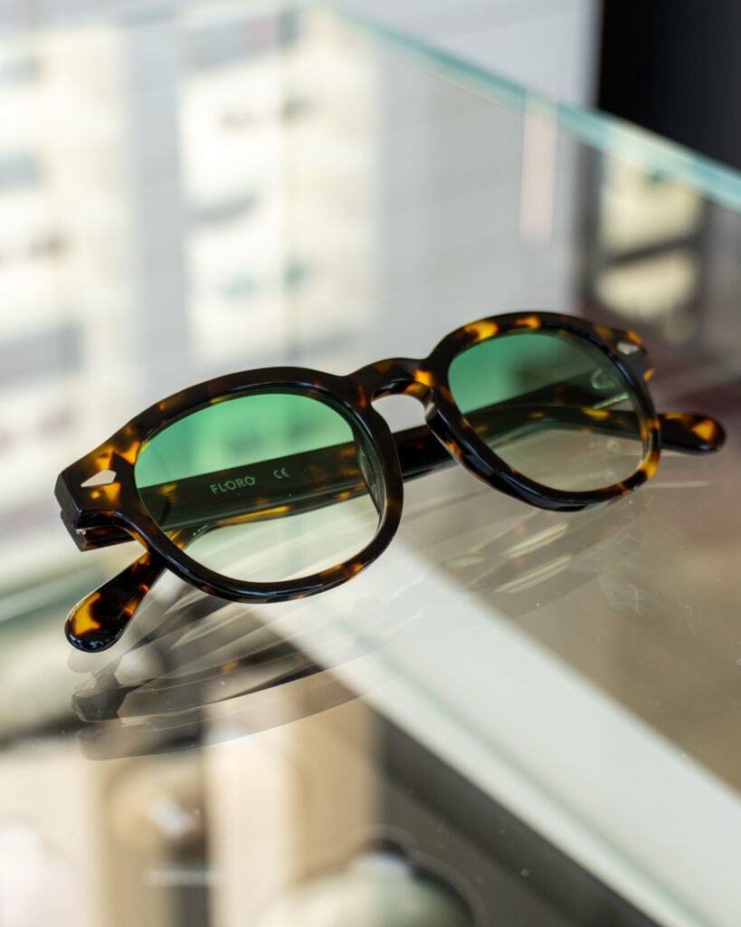 Occhiali da sole Floro in acetato tartaruga con lenti verdi appoggiati su una teca in vetro